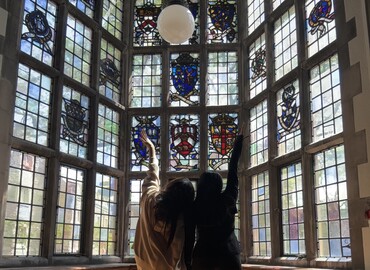 Image of Two Friends Raising their Hands Facing the Stained Glass Window of the Lunch Hall in Trinity College at the University of Toronto. Photo Credits: Kylie Ng